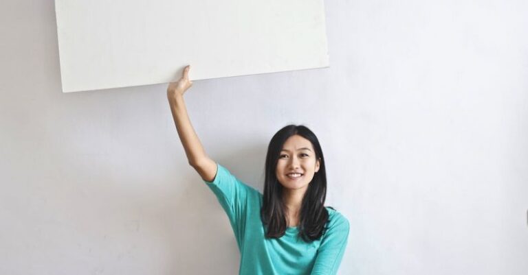 Mortgages - Cheerful Asian woman sitting cross legged on floor against white wall in empty apartment and showing white blank banner