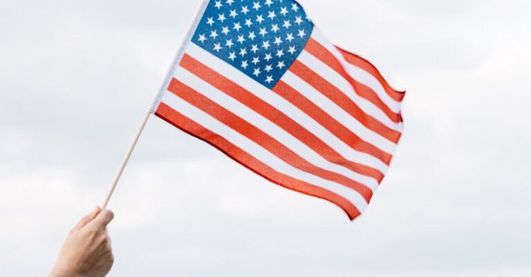 Equity - Back view of crop anonymous patriot showing pointed stick with national flag of America as symbol of heroism and prowess on Independence Day under cloudy sky on green field