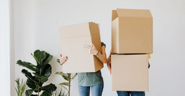 Mortgage - Unrecognizable couple wearing jeans standing carrying stacked carton boxes out of apartment during renovation on daytime